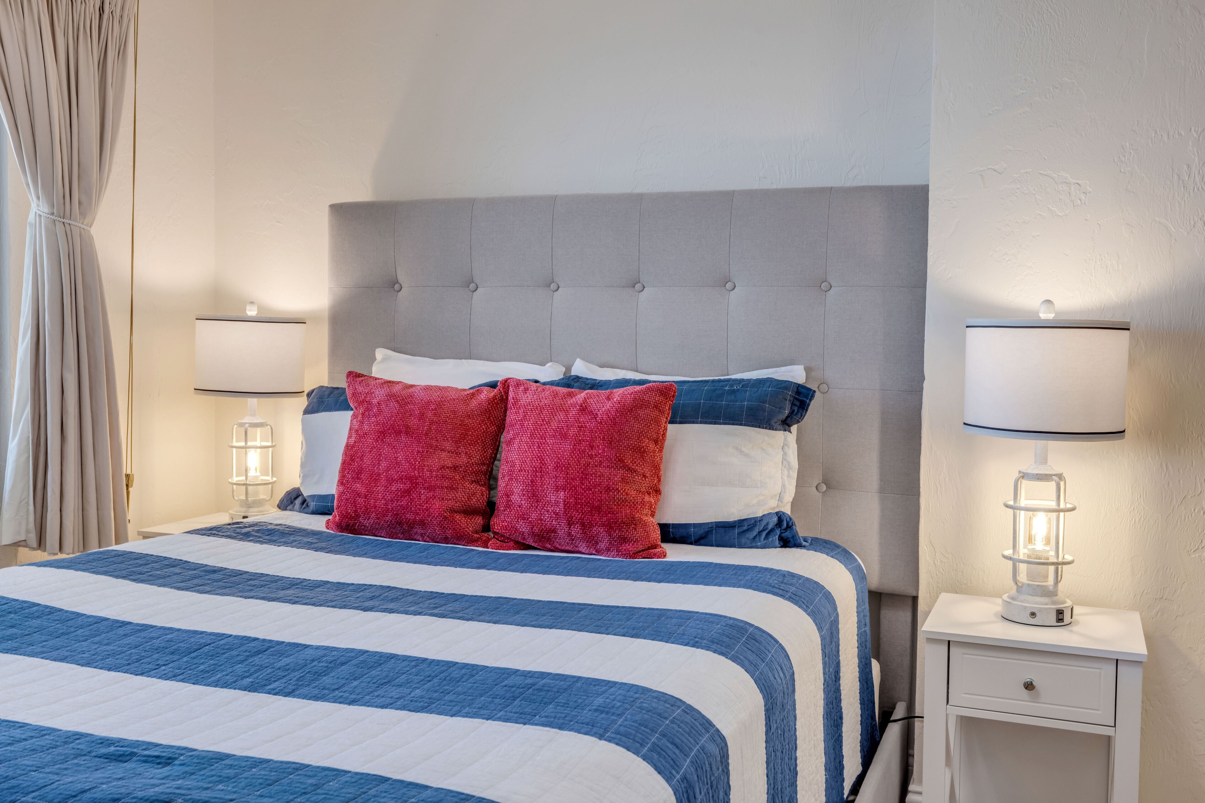 Un lit bien fait, avec une couvre-lit à rayures, une tête de lit rembourrée de couleur grise et deux lampes de chevet.