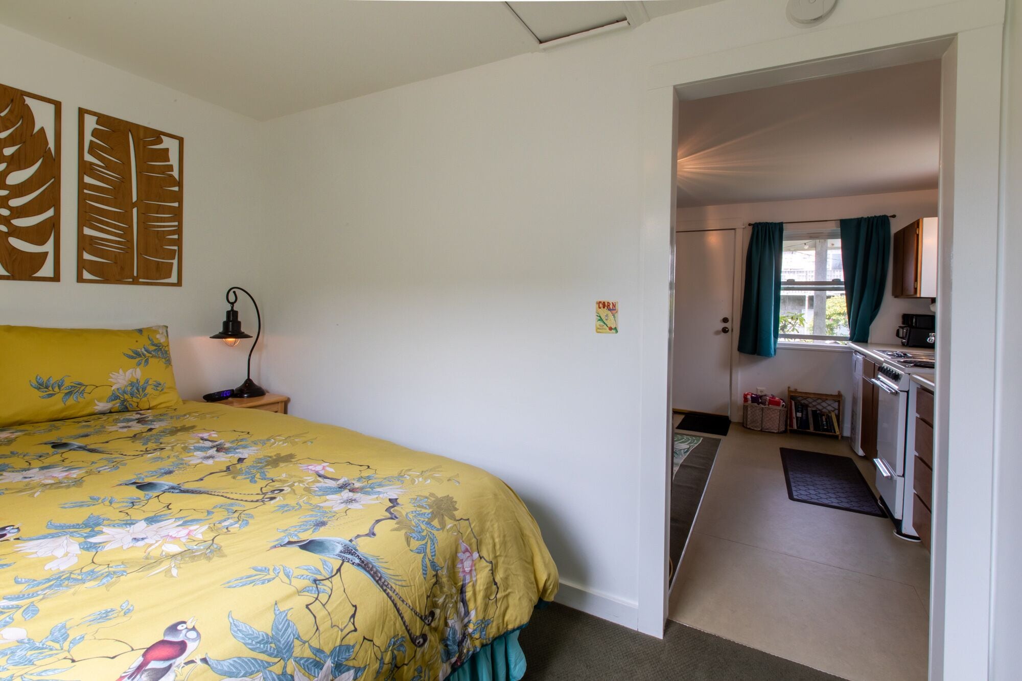Une chambre à coucher avec un lit, une table de chevet et une lampe. On aperçoit une cuisine à travers une porte ouverte.
