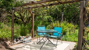 Una pérgola de madera con una mesa de vidrio y dos sillas azules en un jardín de follaje exuberante.