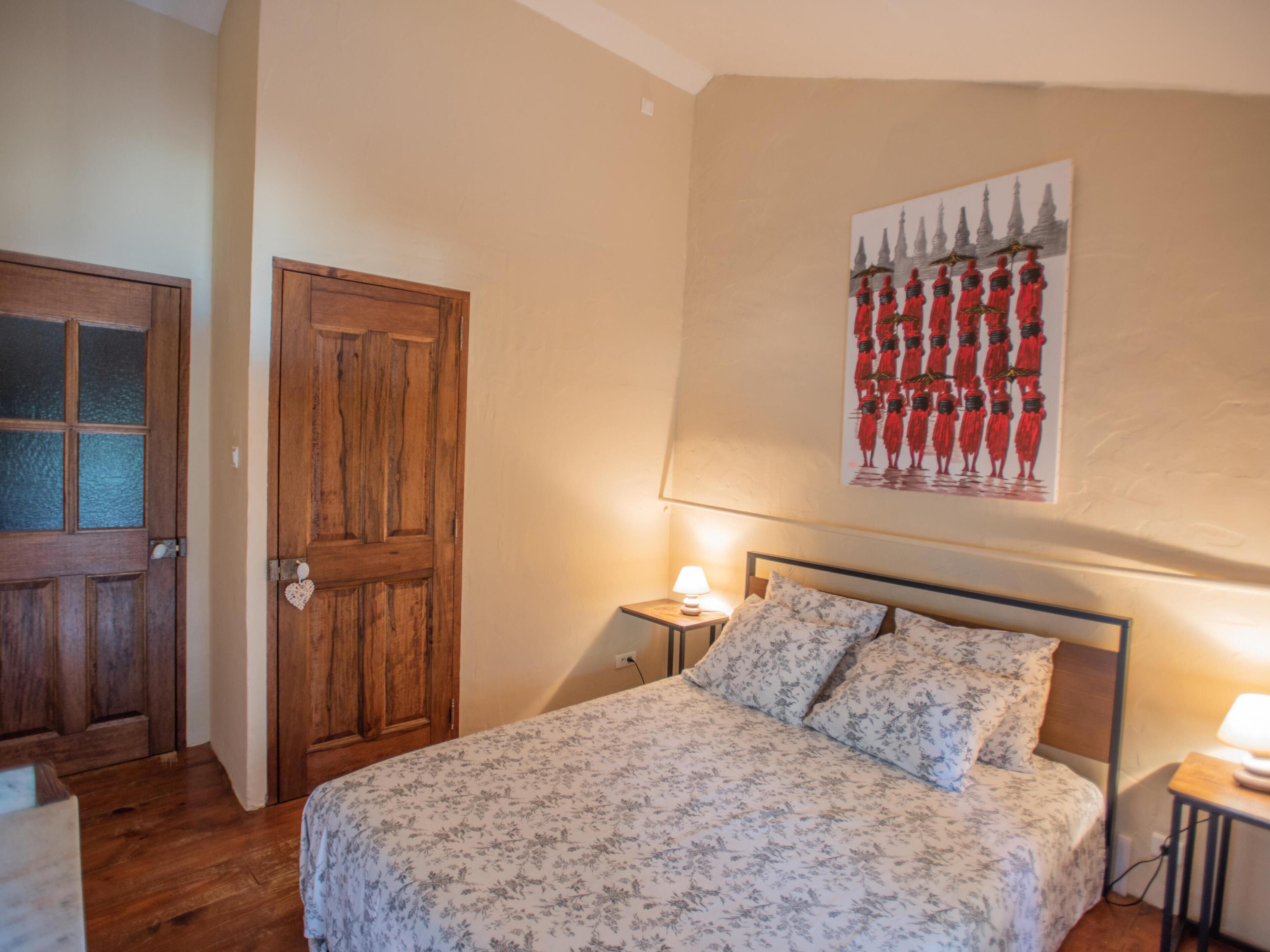 A bedroom with a wooden door, a bed with floral bedding, a nightstand with a lamp, and a wall-mounted painting of figures in red.