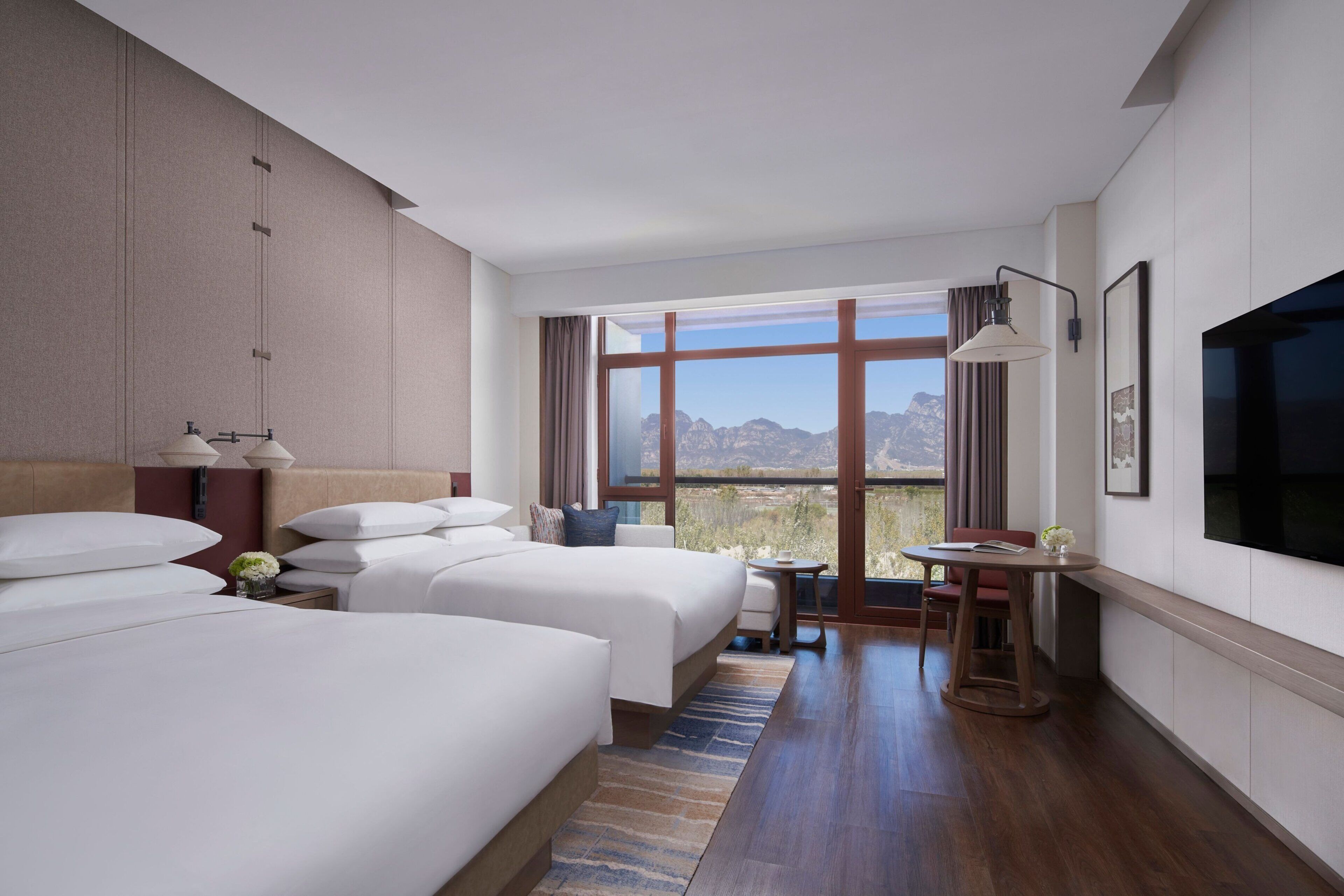 A hotel room with two beds, a desk, a chair, a TV, and a large window with a mountain view.