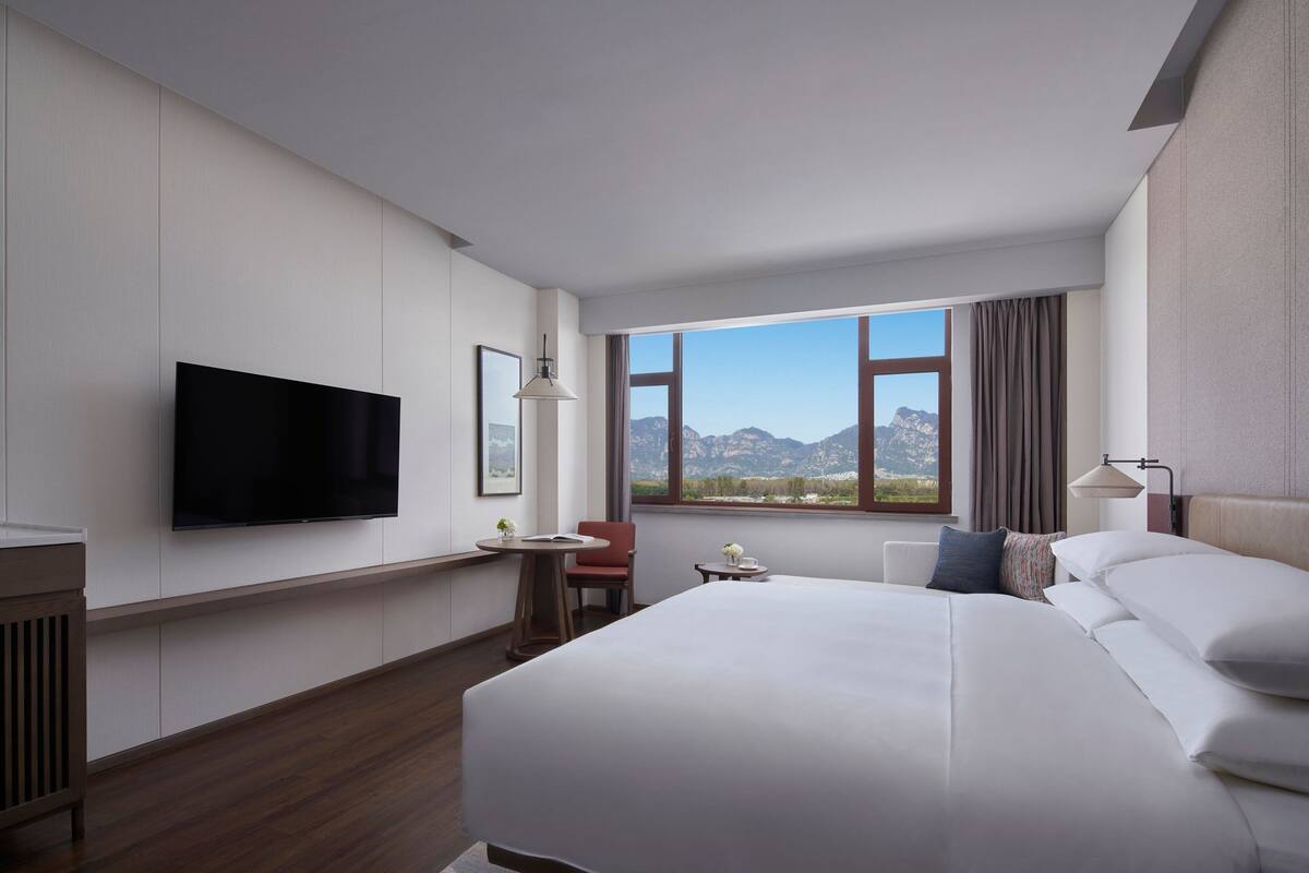A modern hotel room with a large bed, a flat-screen TV, a desk, and a window with a mountain view.