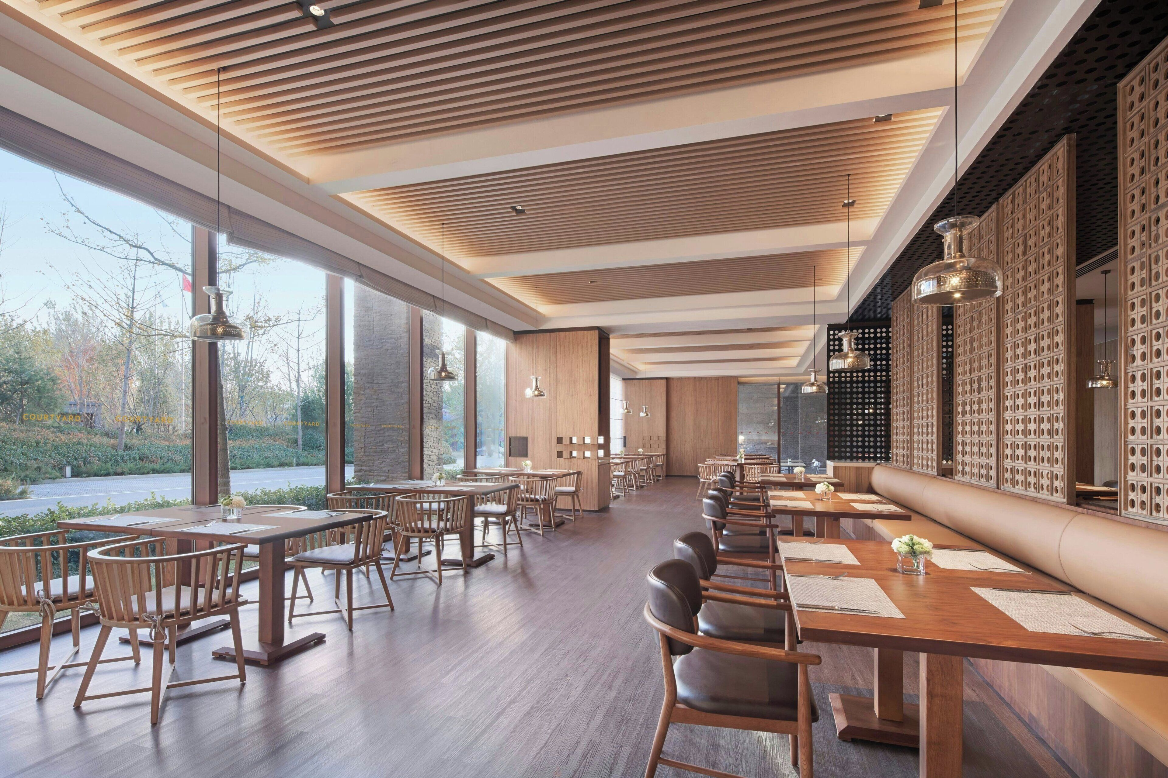 A modern restaurant with wooden tables and chairs, large windows, and a high ceiling with wooden slats.