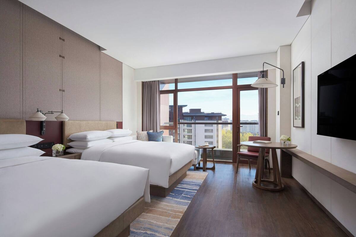 A hotel room with two beds, a TV, a desk, and a large window with a city view.
