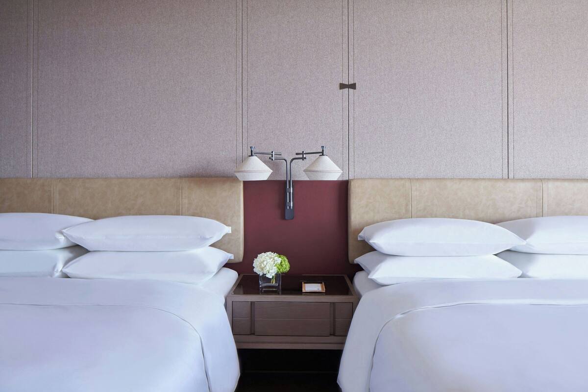 A hotel room with two beds, a nightstand with a vase of flowers, and a wall-mounted lamp.