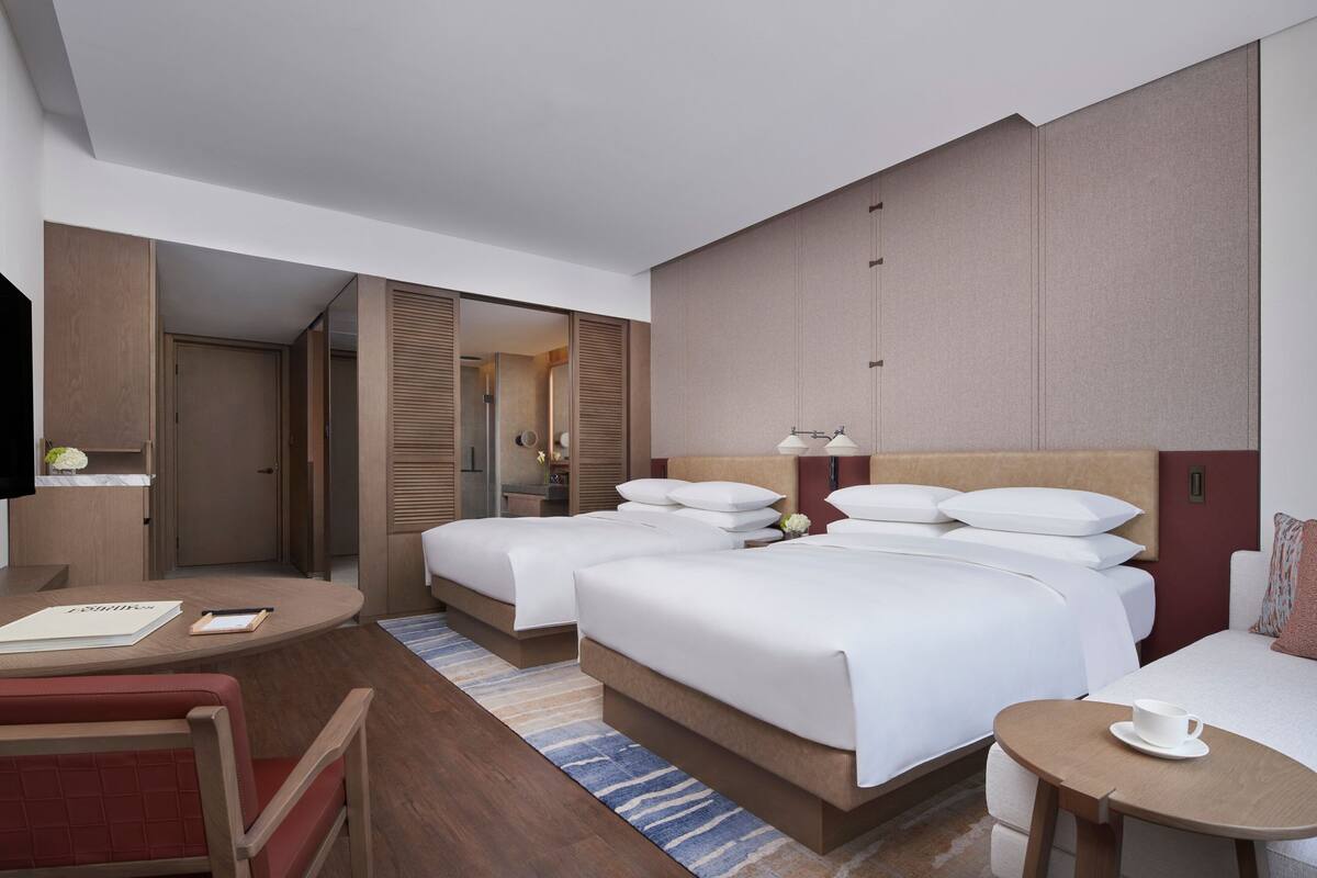 A hotel room with two beds, a desk, a chair, and a small table with a cup.