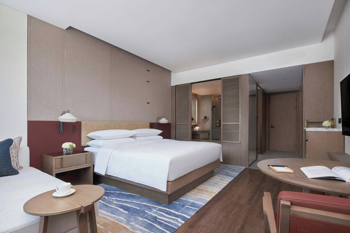 A hotel room with a bed, bedside table, lamp, and a small table with a cup.