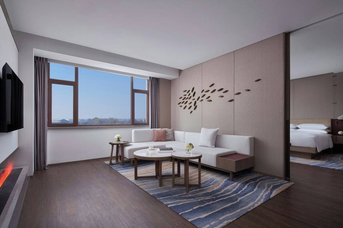 A modern hotel room with a large window, a sofa, two small tables, and a decorative wall panel.