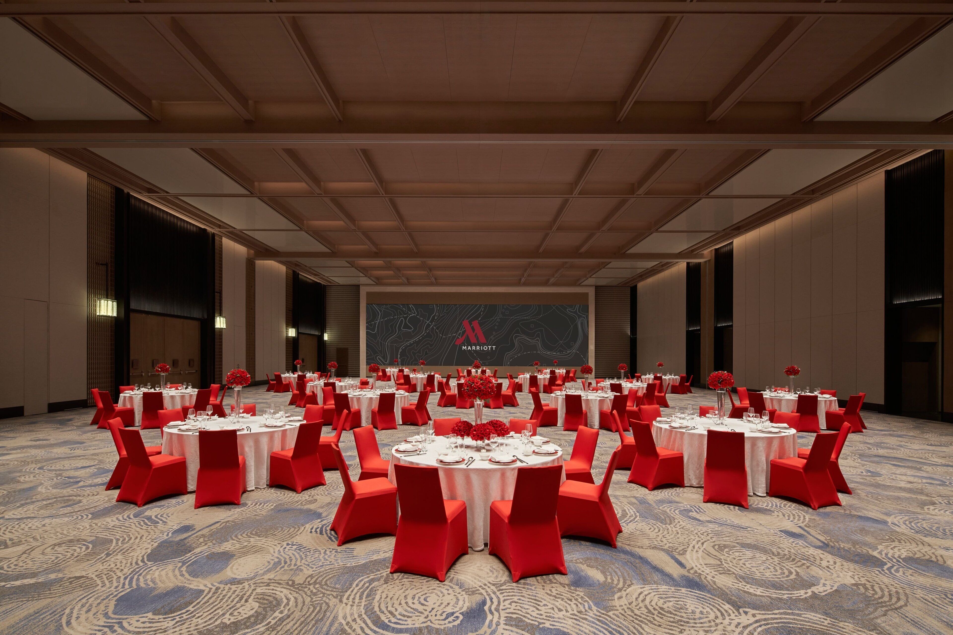 A large conference room with multiple round tables, each set with white tablecloths and red chairs, and a large screen displaying the Marriott logo.