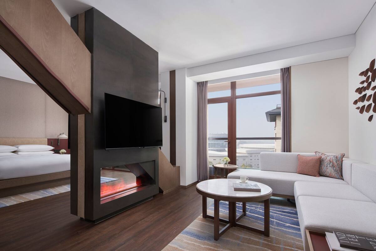 A modern hotel room with a flat-screen TV, a fireplace, a sofa, and a balcony with a view.
