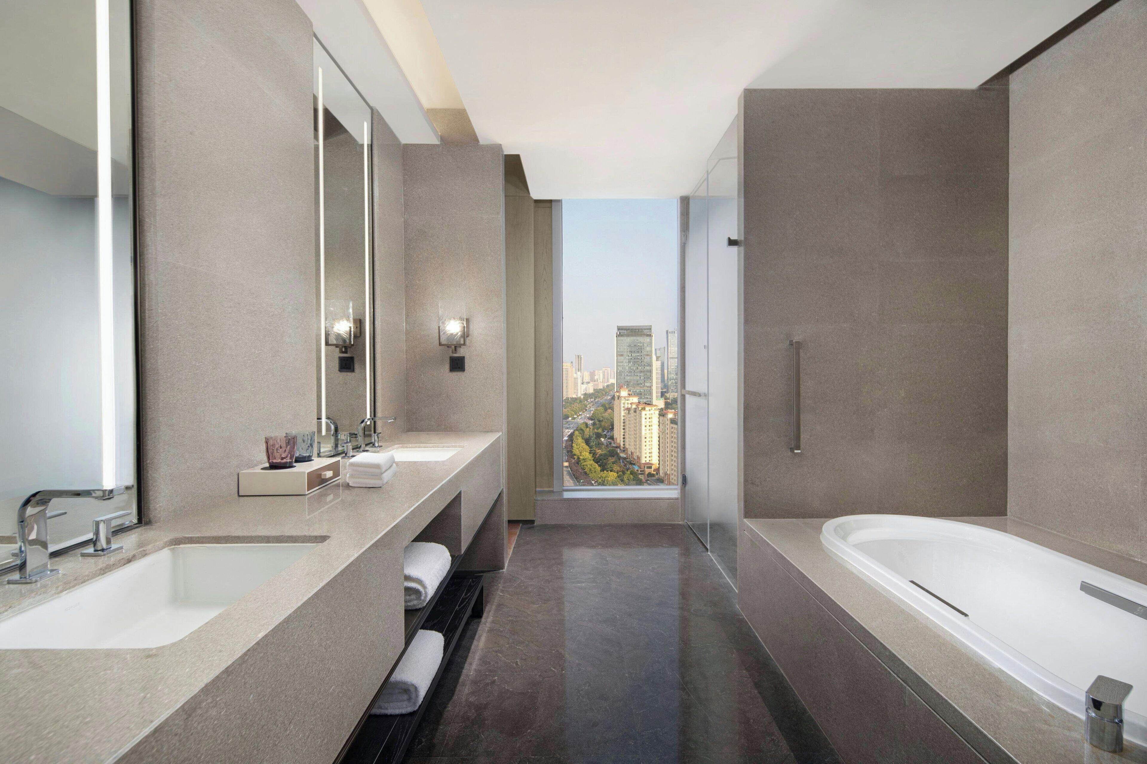 A modern bathroom with a large bathtub, a double sink vanity, a mirror, and a view of the cityscape.