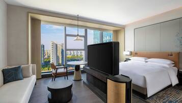 Una habitación de hotel moderna con una cama grande, televisión, sofá, mesa de comedor y vistas a la ciudad.