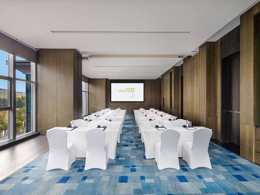 Un salón de conferencias con filas de mesas y sillas blancas, una pantalla grande al frente y ventanas amplias con vista a la ciudad.