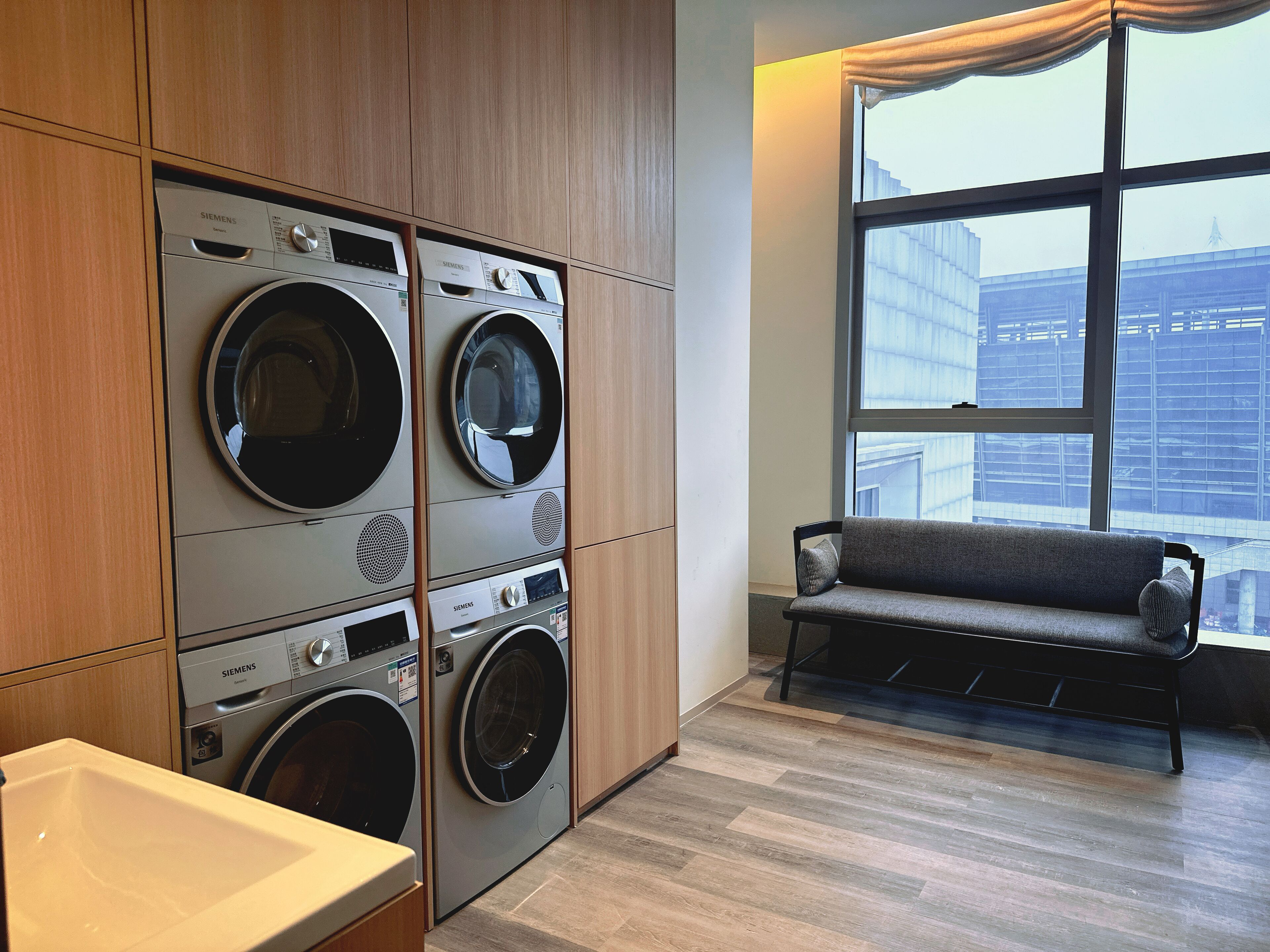 A laundry room with two stacked front-loading washers and dryers, a bench, and a large window with a view of a building.