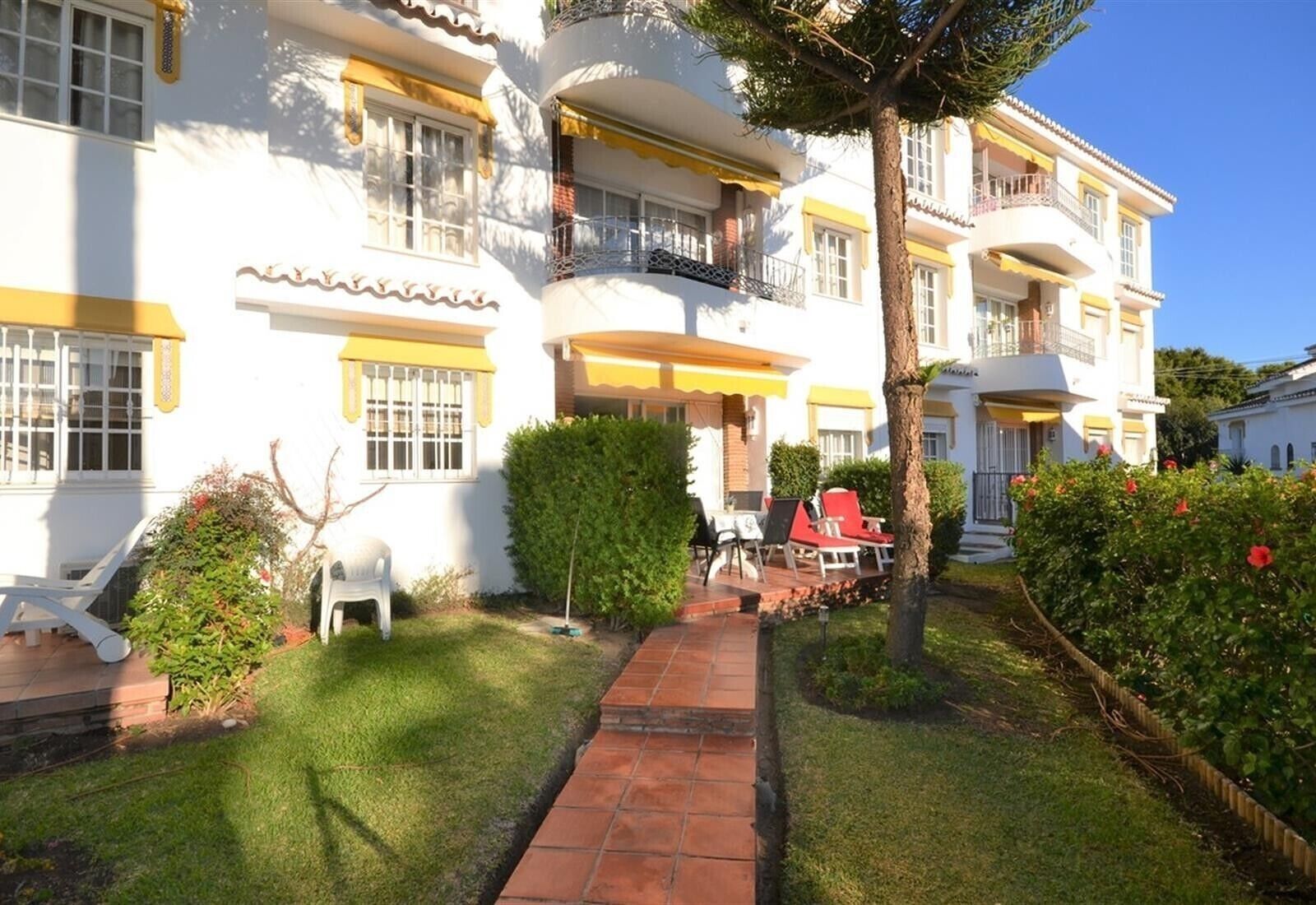 Un edificio de varios pisos con balcones, un camino pavimentado y mobiliario exterior.