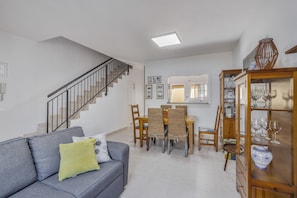 Uma sala de estar com um sofá cinzento, uma escada, uma área de jantar com mesa e cadeiras, e um armário com copos.
