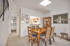 Uma área de refeições com uma mesa e cadeiras de madeira, uma cozinha ao fundo e uma pintura de paisagem urbana na parede.