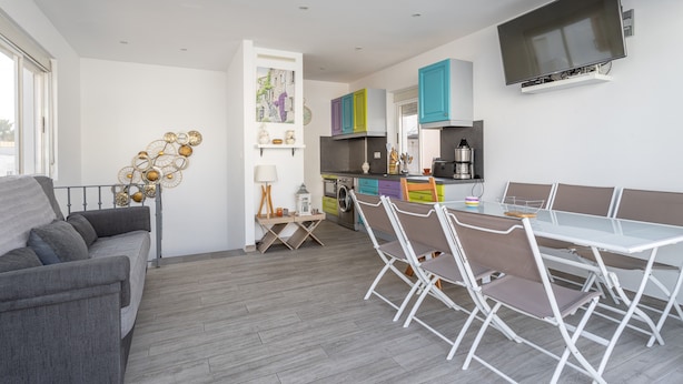 Un espacio de estar moderno con sofá, mesa de comedor, sillas, zona de cocina y televisor montado en la pared.