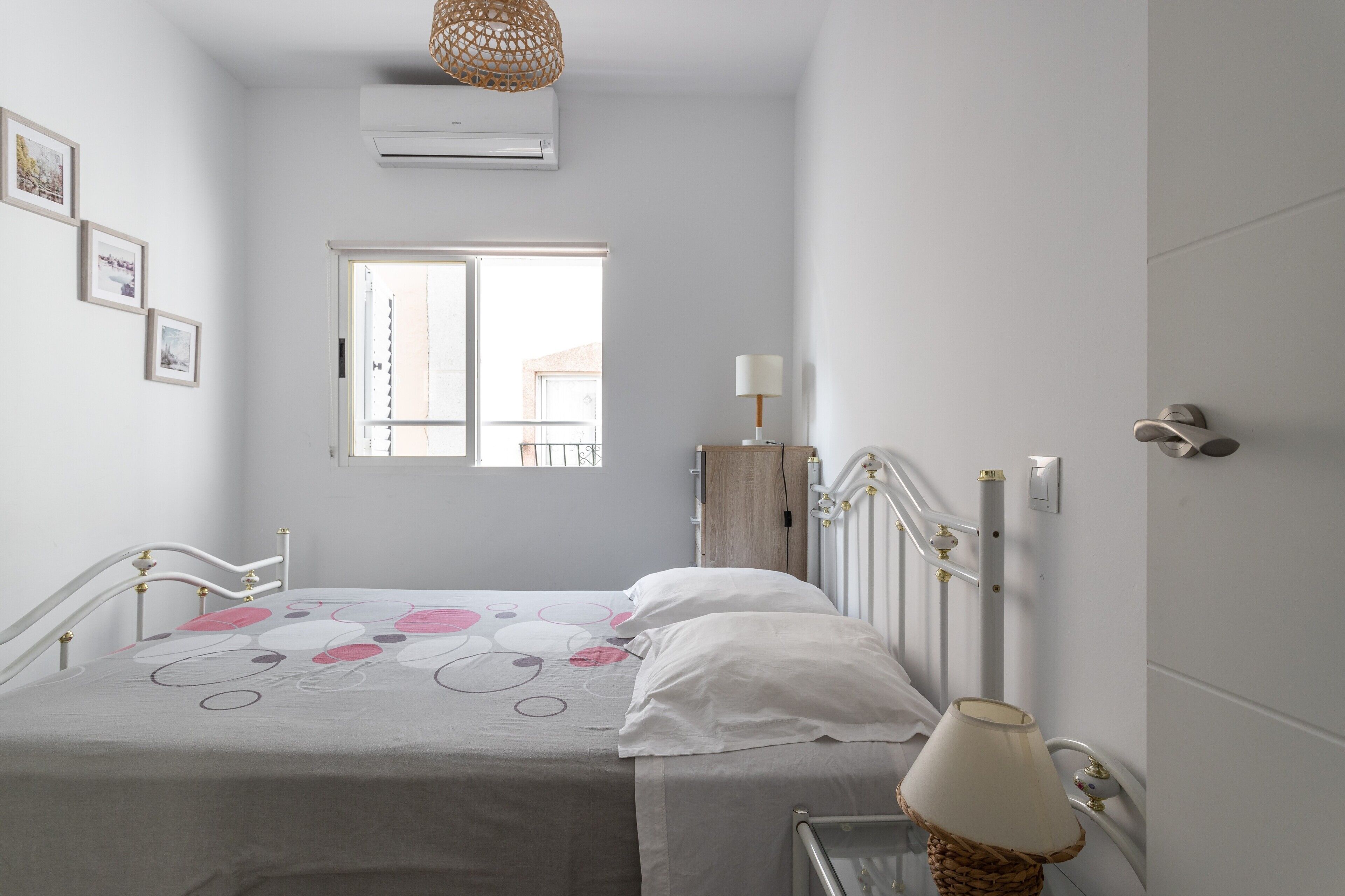 Une chambre à coucher comprenant un lit, une table de chevet, une lampe, une fenêtre, un appareil de climatisation et un interrupteur fixé au mur.