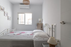 Ein Schlafzimmer mit Bett, Nachttisch, Lampe, Fenster, Klimaanlage und Wandleuchte.