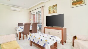 Habitación de hotel con mesa de comedor, sillas, televisor, cama y ventana con cortinas.