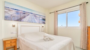 Un dormitorio con cama, mesita de noche, un cuadro, ventana y cortinas.