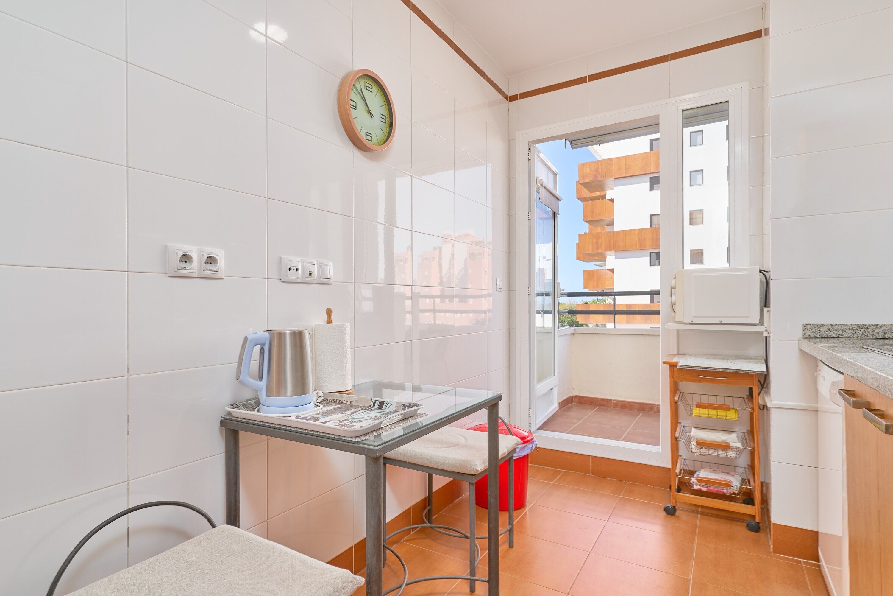 Une cuisine avec des carreaux blancs, une horloge, une machine à café, une petite table et une vue sur un balcon donnant sur des immeubles.