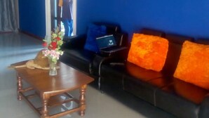 A reception area with a blue wall, a wooden coffee table with a vase of flowers, a black sofa with orange cushions, and a framed certificate on the wall. - Muroy Executive Lodge (Kitwe)