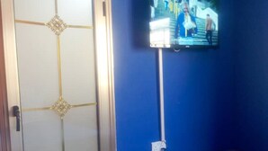 A room with a blue wall, a television mounted on the wall, a door with a decorative pattern, and a small gray cabinet. - Muroy Executive Lodge (Kitwe)