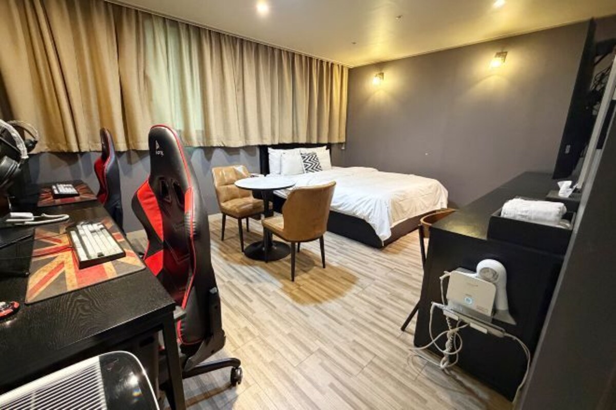 A hotel room with a bed, a desk with a computer, a gaming chair, a small round table, and a hairdryer on a stand.