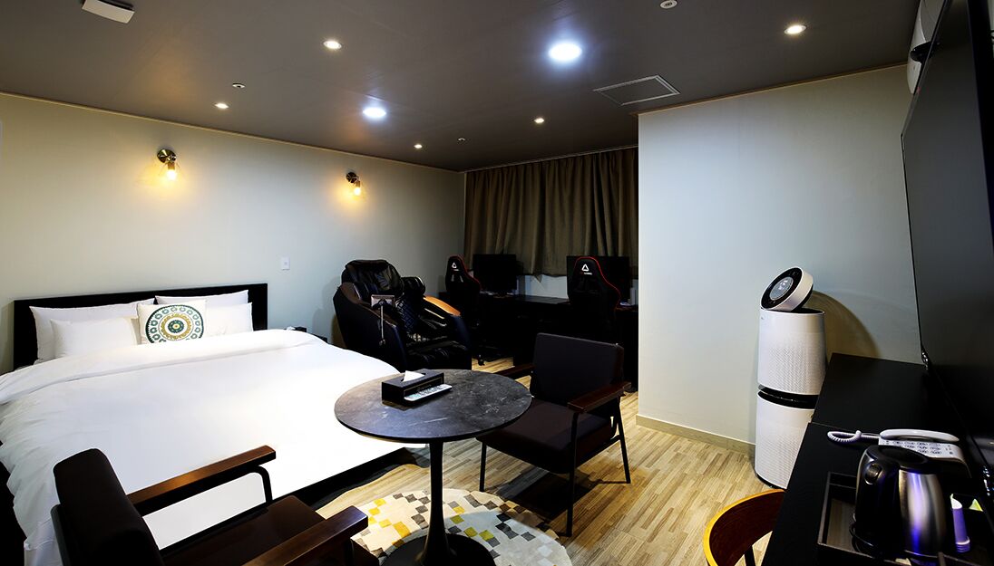 A hotel room with a bed, a desk with a computer, a chair, a small table, and a coffee machine.