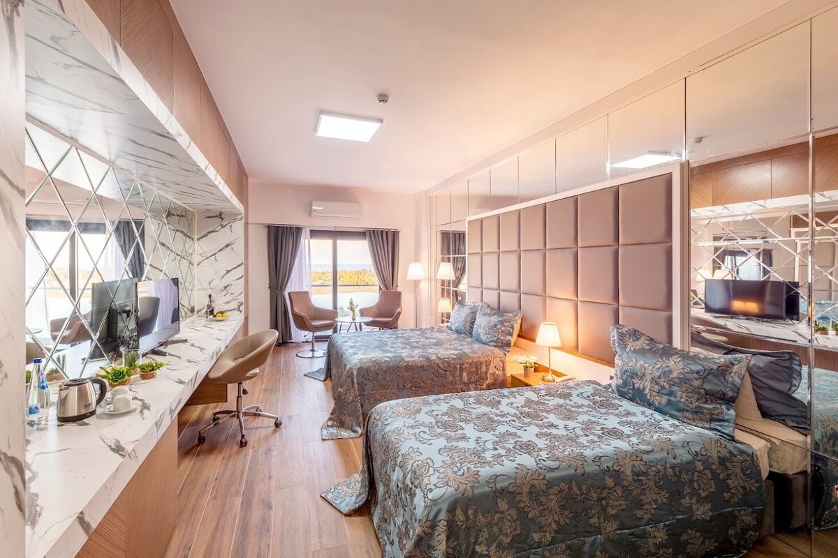 A hotel room with two beds, a desk with a computer, a TV, and a seating area with chairs.