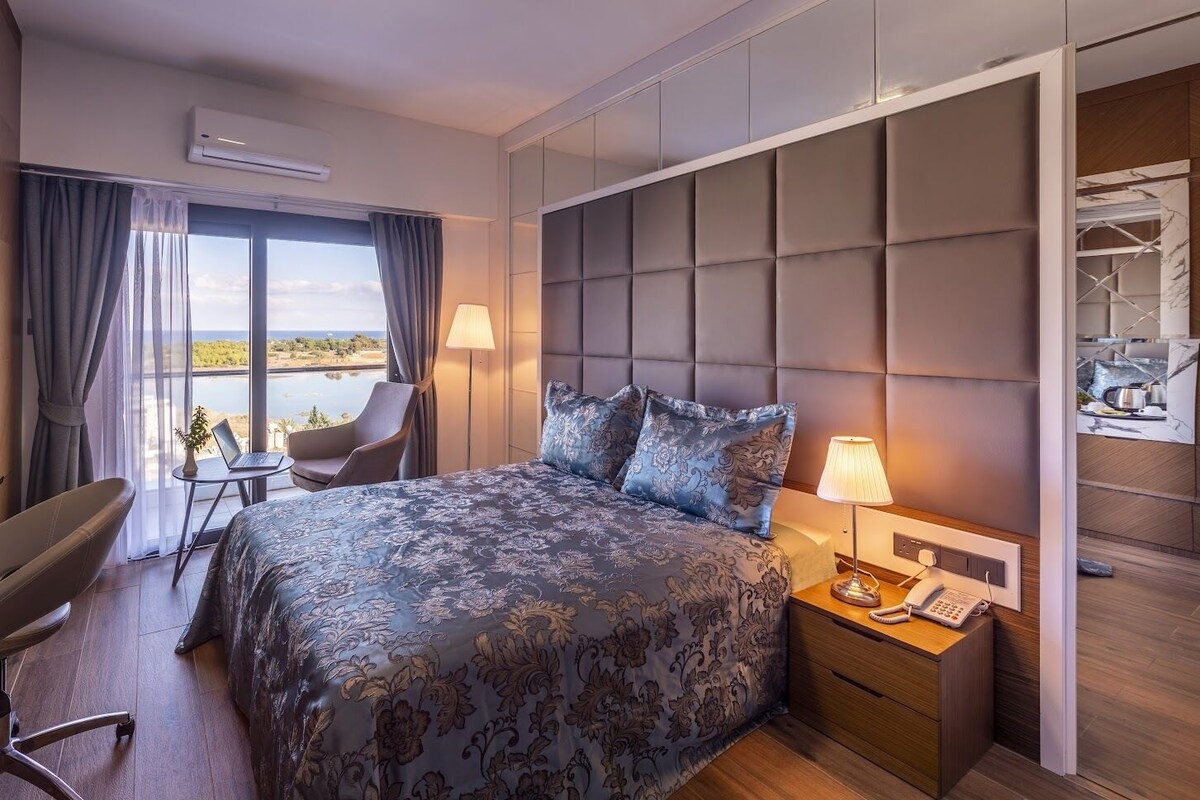 A modern hotel room with a large bed, a desk, a chair, and a view of the outdoors.
