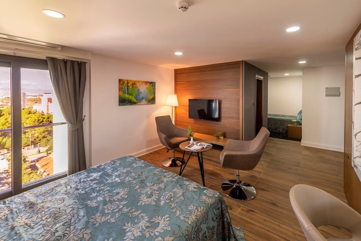A hotel room with a bed, a desk, two chairs, a TV, and a view of the city.