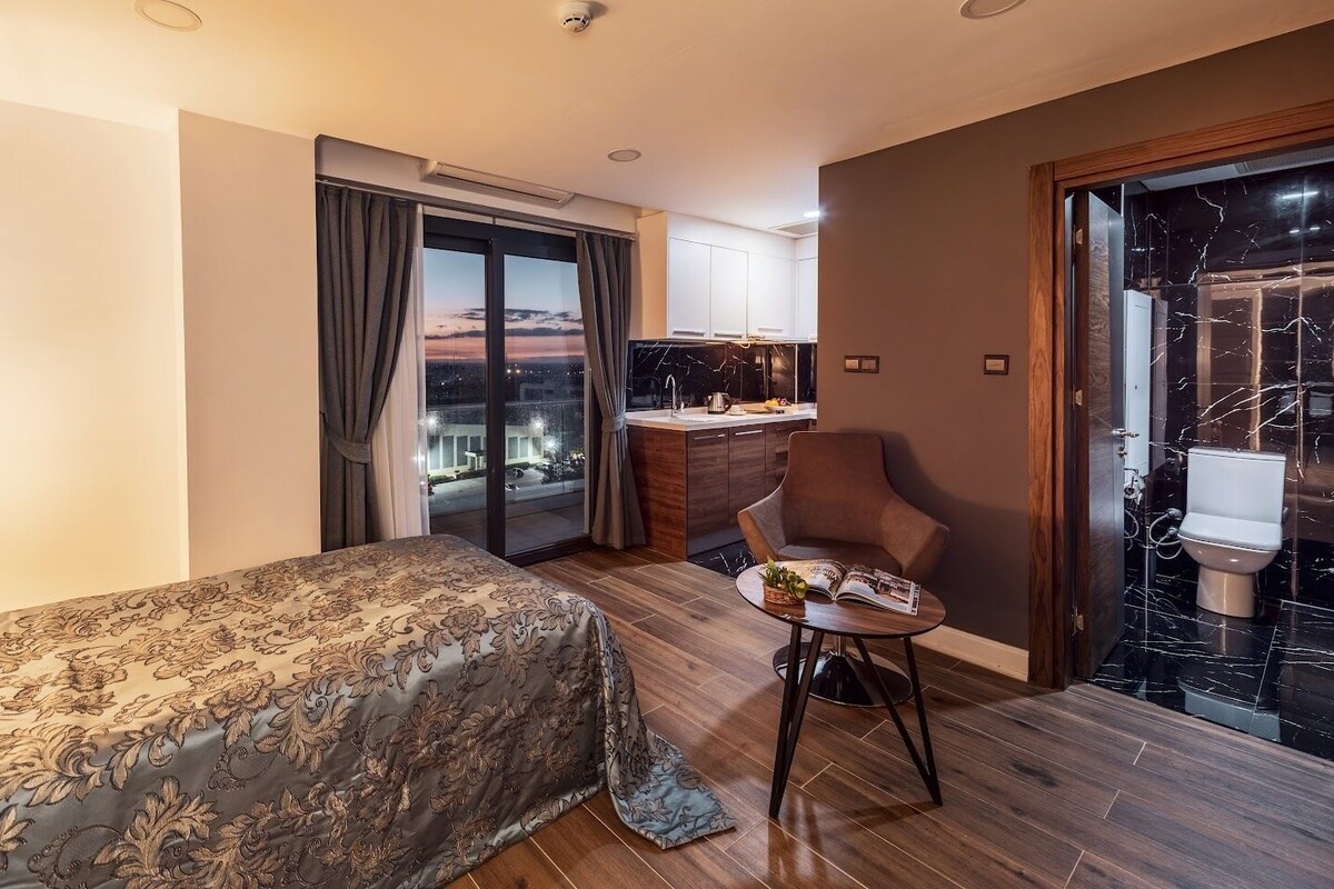 A hotel room with a bed, a small table with a magazine, a chair, a view of the city at night, and a bathroom with a marble floor.