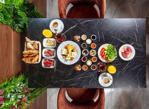 A breakfast table with various foods and drinks, surrounded by chairs and plants. - Novel Centre Point Hotel (Famagusta)
