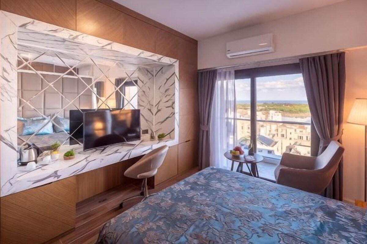 A hotel room with a bed, a TV, a small table with a plant, a chair, and a window with a view.