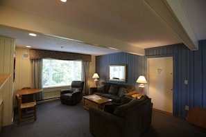 Una sala de estar amoblada con sofá, sillón y una mesa, una ventana con cortinas y una puerta con un letrero.