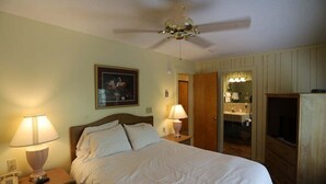 Quarto de hotel com uma cama, duas luminárias de cabeceira, ventoinha no teto, um quadro na parede, um espelho e uma televisão.