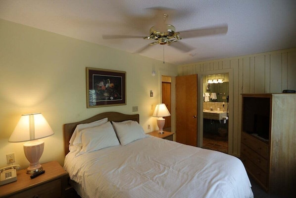 A hotel room with a bed, two bedside lamps, a ceiling fan, a painting on the wall, a mirror, and a TV.