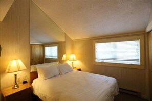 Una habitación de hotel con cama, dos lámparas de noche, una ventana con persianas y un espejo que refleja la habitación.