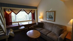 A room with a brown sofa, a wooden table, a window with curtains, a framed picture on the wall, and a lamp. - Inns of Waterville Valley Loft Unit (Waterville Valley)