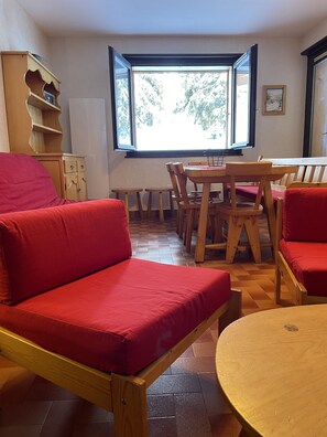 Une pièce comprenant une table à manger en bois, des chaises à coussins rouges, un canapé en bois et une fenêtre donnant sur des arbres.