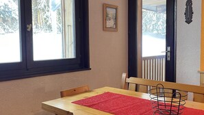 En matplats med ett träbord och stolar, en röd matta och en korg av metall. Det finns två fönster med svarta ramar och en dörr med en svart ram. En tavla hänger på väggen.