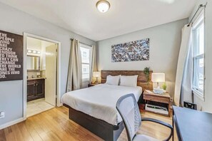 Camera d'albergo moderna con un letto, comodini, una sedia, una scrivania e un bagno parzialmente visibile attraverso una porta aperta.