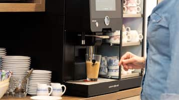 Eine Kaffeemaschine füllt ein Getränk in ein Glas, während sich Geschirrgestelle mit Tellern und Tassen auf der Theke befinden.