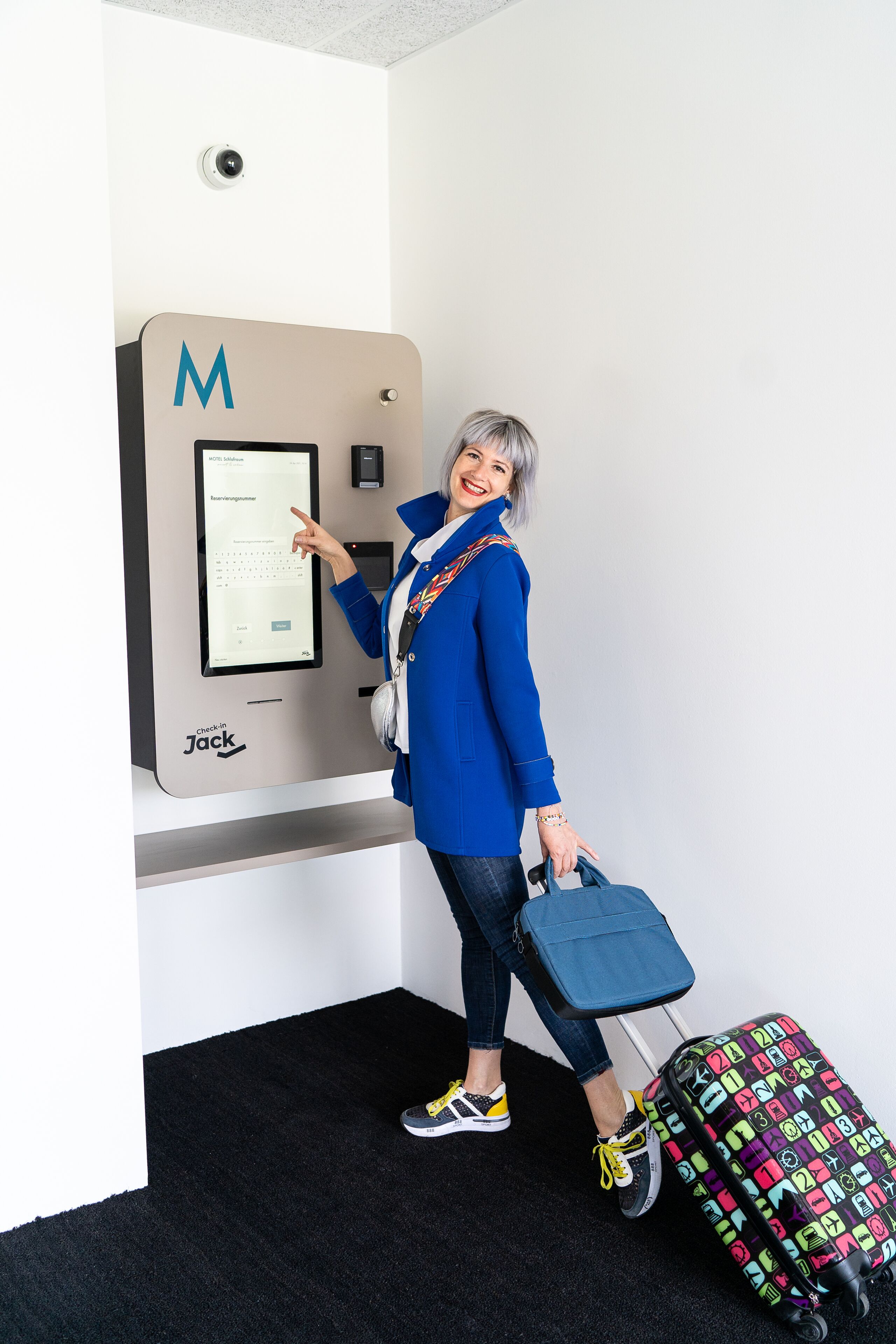 Una persona utilizza una postazione self check-in etichettata 'Jack' in una stanza con tappeto nero e parete bianca.