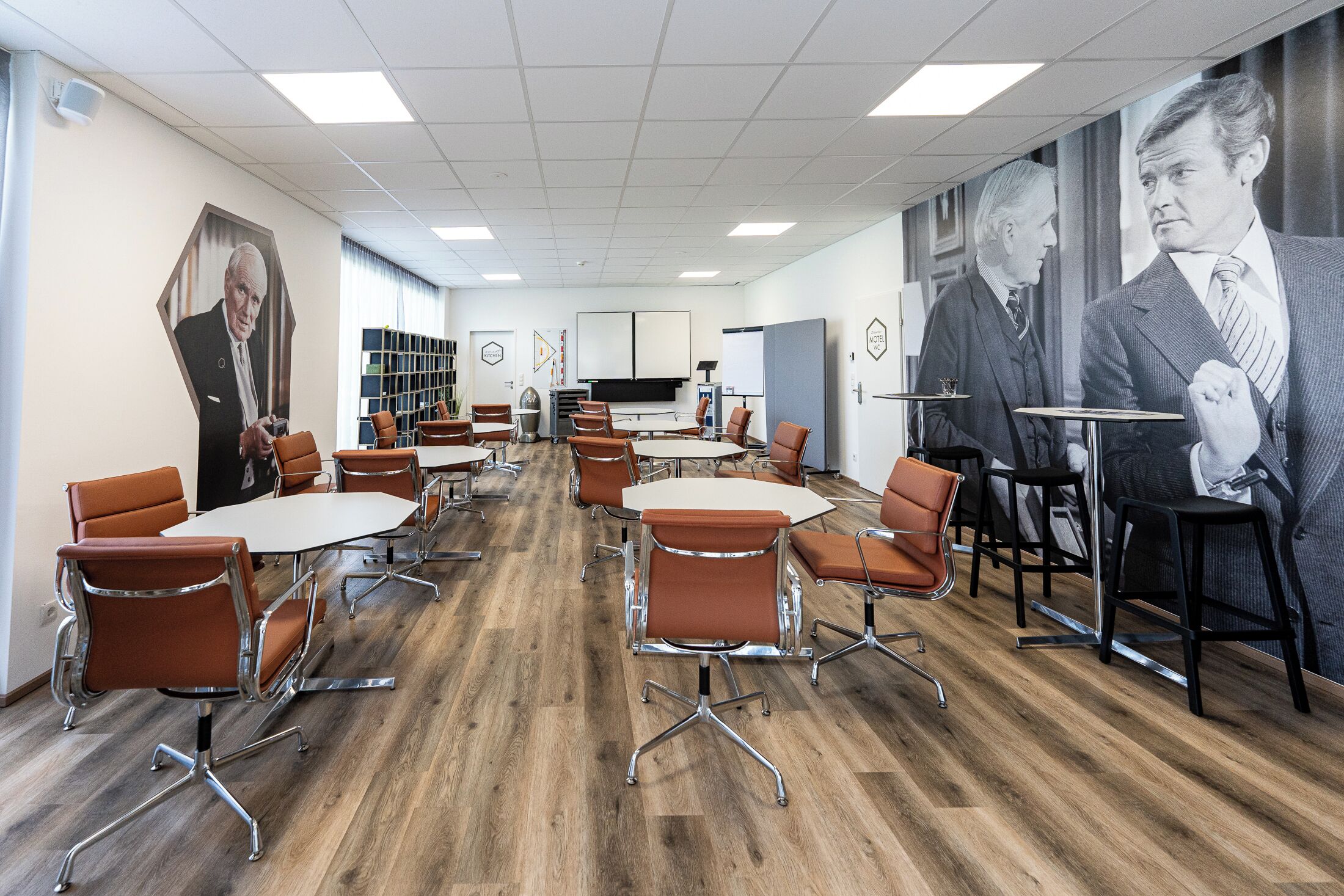 A conference room with round tables and orange chairs, a large screen, and bookshelves in the background.