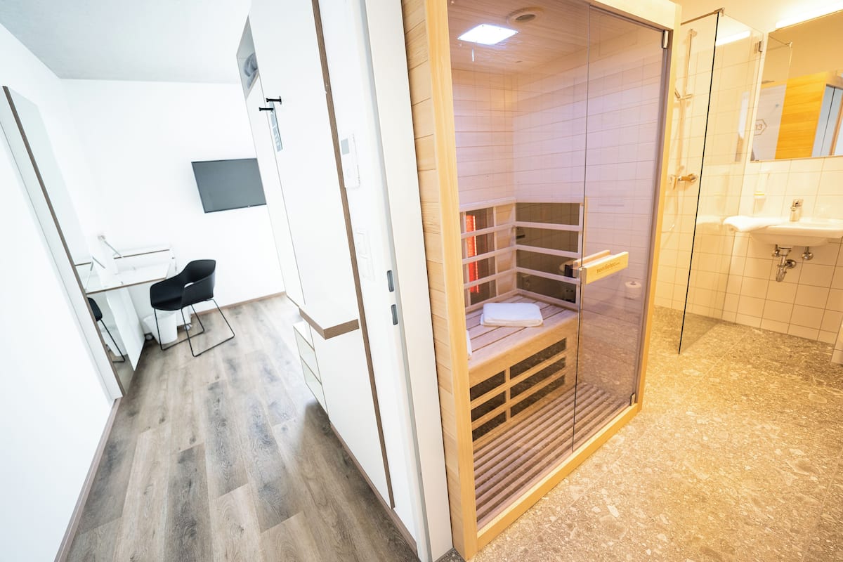 Une chambre d’hôtel moderne avec un plancher en bois, un bureau avec une chaise, une douche séparée par une paroi de verre et une salle de bain équipée d’un lavabo et d’un miroir.