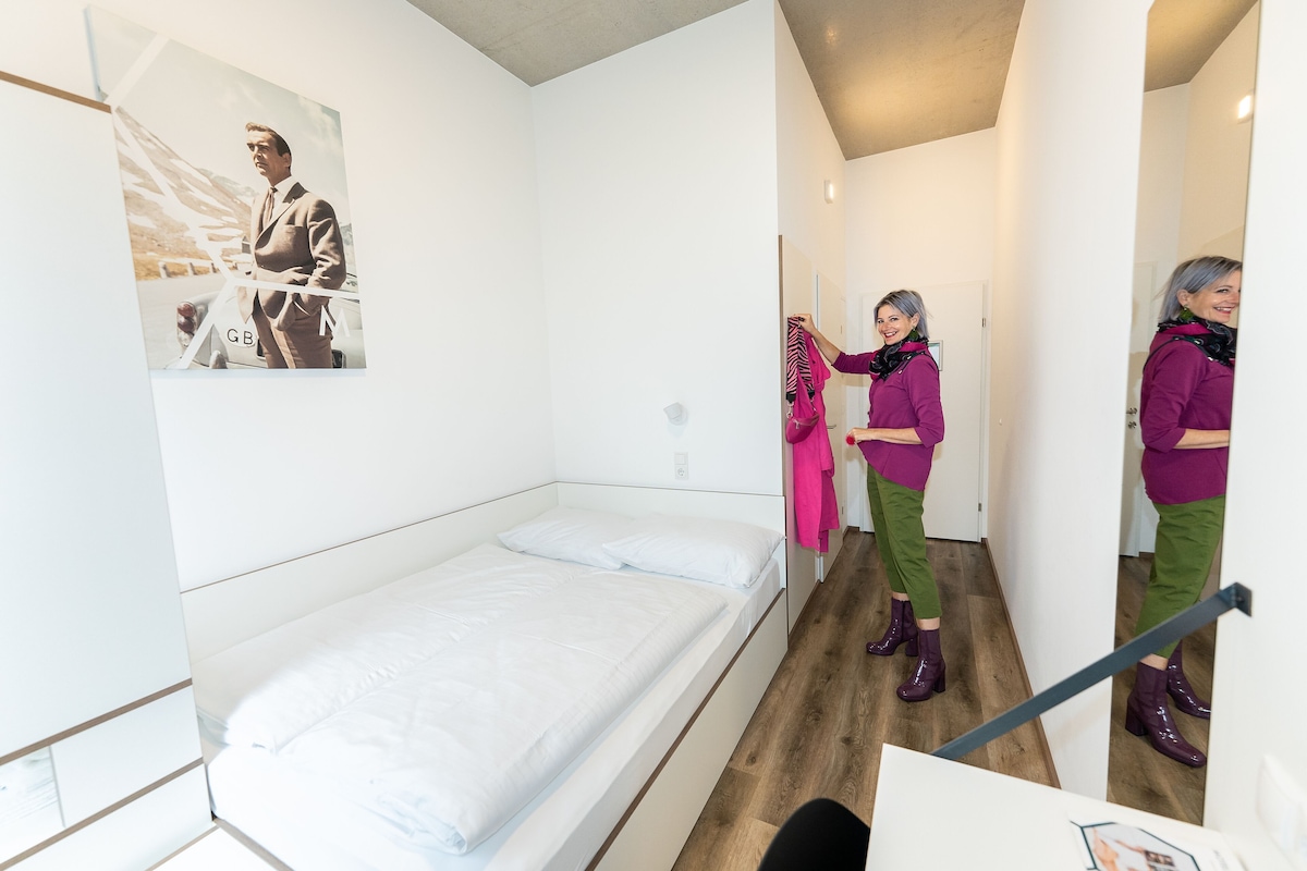 Habitación individual con cama, piso de madera, pared blanca y un cuadro enmarcado.